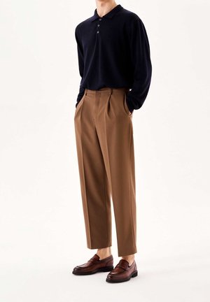 Polo à manches longues bleu marine, pantalons plissés beiges et mocassins marron. La tenue présente une coupe classique avec des textures lisses et un design simple.