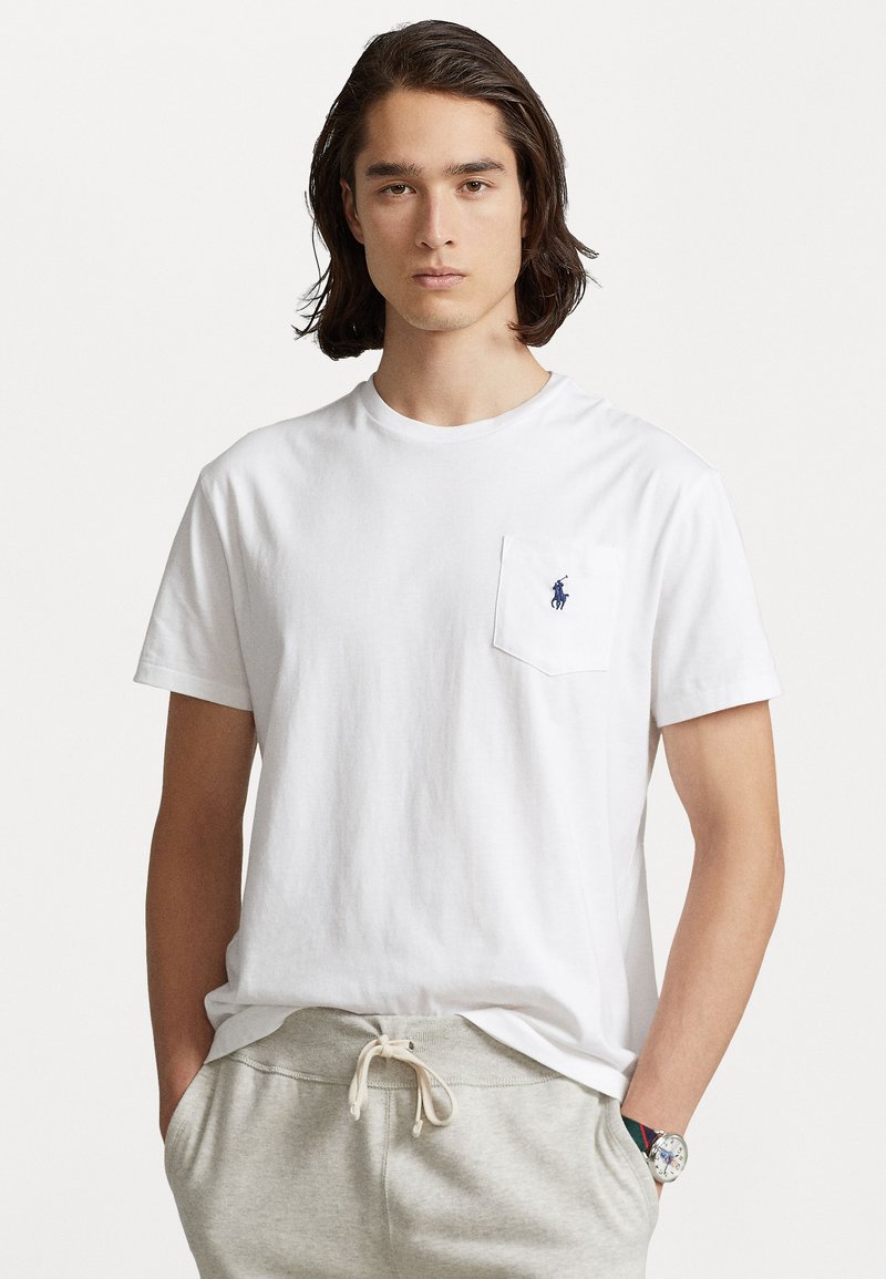 Tricou alb din bumbac cu guler rotund și mâneci scurte, având o buzunar pe pieptul stâng și un mic logo albastru pe buzunar.