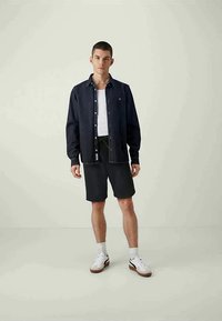 Jovem vestido com uma jaqueta azul escura sobre uma t-shirt branca, calções pretos, meias brancas e ténis brancos, de pé contra um fundo claro.