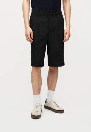 Muž nosi čierne na mieru šité šortky po kolená, biele ponožky a béžové tenisky, stojaci pred jednofarebným svetlým pozadím.