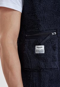 Marinblå fleeceväst med en texturerad yta, som har en dragkedjeficka och en etikett. Materialet verkar mjukt och varmt.
