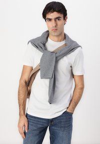 Un sweat-shirt gris noué autour du cou sur un t-shirt blanc, associé à un jean bleu. Fond neutre, mettant en valeur les couches et les couleurs de la tenue.