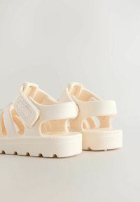 Beige rubber sandals with open design, velcro strap, thick sole, and textured bottom for grip. Features soft edges and minimal branding.