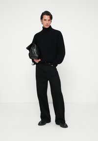 Homme portant un pull col roulé noir et un pantalon large noir, tenant un sac en cuir noir contre un fond blanc uni.