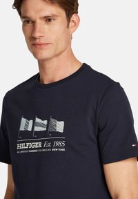 Camiseta de algodón azul marino con un gráfico de tres banderas estilizadas, el nombre de la marca y el año de establecimiento en impresión blanca, cuello redondo y mangas cortas.