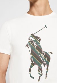 Bílé bavlněné tričko s barevným pruhovaným motivem hráče polo na koni. Design obsahuje zelené, červené a žluté linky.