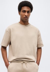 Άνδρας με κοντά σγουρά μαλλιά που φοράει μπεζ φούτερ με κοντά μανίκια και παντελόνι με κορδόνι ταιριαστό, χέρια στις τσέπες, μπροστά σε απλό φωτεινό φόντο.