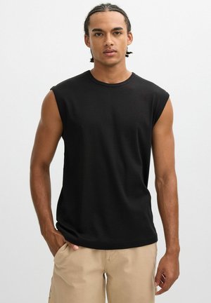 Joven con trenzas, camisa negra sin mangas y pantalones beige, de pie con una mano en el bolsillo sobre un fondo blanco.