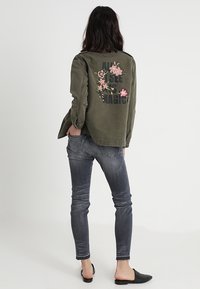 Olivekowa bomberka z kwiatową haftem na plecach, czarny nadruk, zestawiona z szarymi obcisłymi dżinsami i czarnymi wsuwanymi butami.