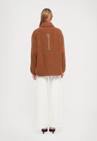 Donna in piedi di spalle, con indosso un giubbotto marrone a trama con il grande numero "1" sulla schiena, pantaloni bianchi a gamba larga e tacchi neri.