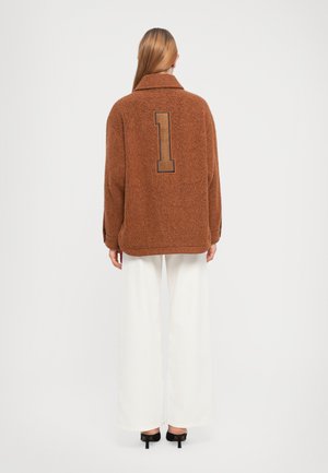 Femme debout de dos, portant une veste texturée marron avec un grand chiffre "1" dans le dos, un pantalon blanc à larges jambes et des escarpins noirs.