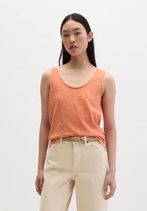 Jeune femme portant un haut orange sans manches rentré dans un pantalon beige taille haute avec une ceinture camel, debout devant un fond blanc uni.
