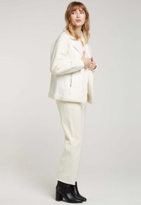 Veste blanche au toucher duveteux avec fermeture éclair sur le côté, portée sur un pantalon crème et des bottines noires à talons. Col simple et coupe ample.