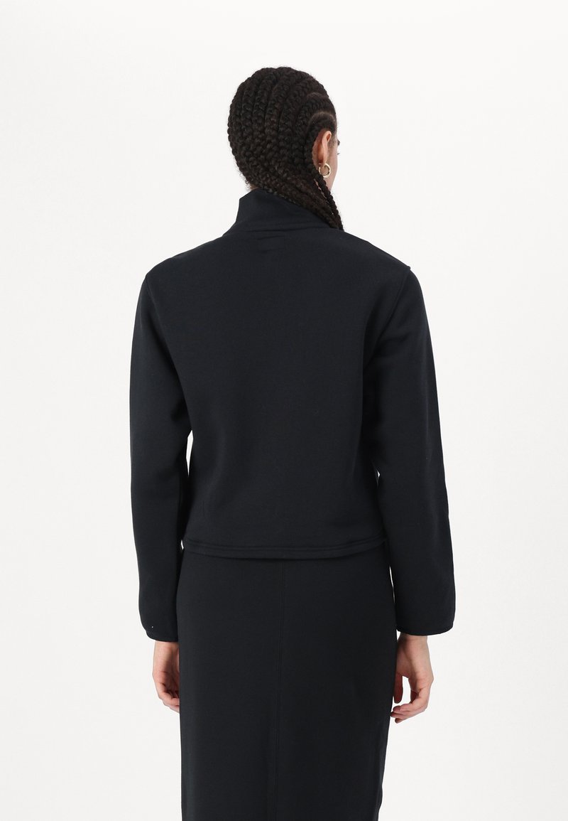 Veste noire ajustée avec un col montant, des manches longues et un tissu texturé, présentée de dos. Design minimaliste, finition lisse.