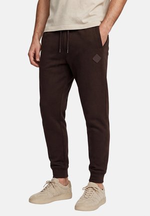 Uomo che indossa pantaloni jogging marrone scuro con tasche laterali e sneakers beige, in piedi con una mano in tasca su sfondo uniforme.