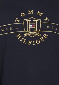 Granatowa bawełniana koszulka z haftowanym złotym logo w kształcie tarczy, zawierającym tekst "TOMMY HILFIGER" oraz "NEW YORK CITY", zdobionym cyframi rzymskimi.