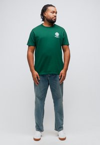 Άντρας με πλεξούδες, φορώντας πράσινο μπλουζάκι με άσπρο έμβλημα, μπλε τζιν και λευκά αθλητικά παπούτσια, στέκεται και κοιτάζει προς τα πλάγια.