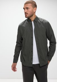 Camisa de botones verde oscuro con un patrón geométrico, mangas largas y cuello clásico, combinada con una camiseta blanca debajo.