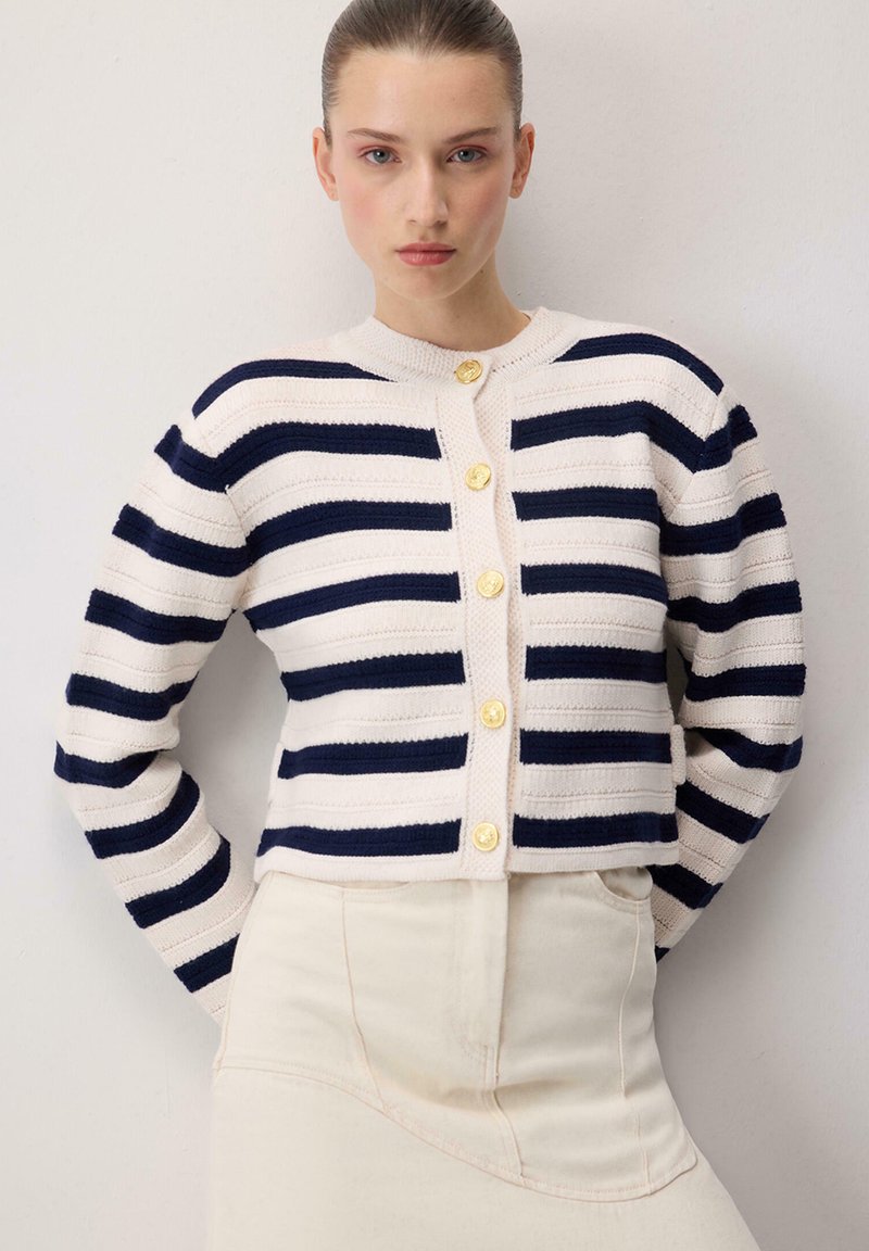 Γυναίκα που φοράει λευκή και ναυτικό μπλε ριγέ ζακέτα με χρυσά κουμπιά και ανοιχτό μπεζ ψηλόμεσο παντελόνι, στέκεται μπροστά σε έναν απλό τοίχο.