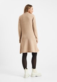 Beiges Strickkleid mit langen Ärmeln und ausgestellter Silhouette, kombiniert mit weißen Knöchelstiefeln. Glatte Textur und knielange Passform.