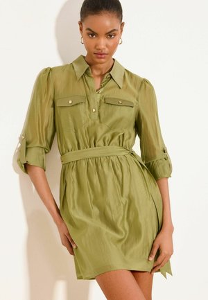 Kobieta ubrana w oliwkową sukienkę z paskiem, bufkami na rękawach, zapinanymi na guziki kieszeniami na piersi oraz złotymi kolczykami kołami na jednolitym białym tle.