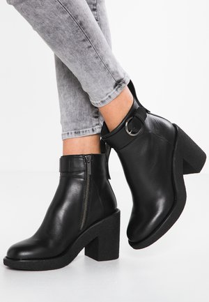 Bottes noires en cuir douce avec un talon épais texturé, zip latéral et un détail en sangle avec une boucle métallique circulaire.