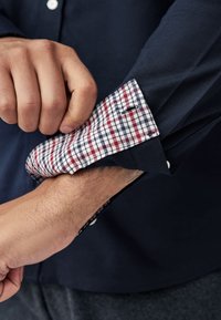 Camicia blu navy con polsino a quadri in rosso, bianco e blu navy. Polsino abbottonato con cuciture visibili e una texture liscia, che lascia intravedere un interno scuro.