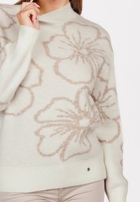 Femme portant un pull crème avec de grands motifs floraux marron clair, tenant le col près de son visage, associé à un pantalon beige clair.
