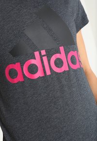 Zbliżenie na osobę noszącą ciemnoszary t-shirt Adidas z czarnym logo marki i jasnoróżowym tekstem na klatce piersiowej.
