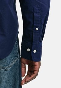 Camicia blu navy con polsini abbottonati e bottoni bianchi. Il materiale sembra liscio con una leggera lucentezza, abbinata a jeans di denim.