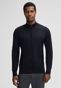 Granatowy kardigan na zamek z wysokim kołnierzem, długimi rękawami i prążkowanymi mankietami. Wąski krój i gładka, lekka tekstura.