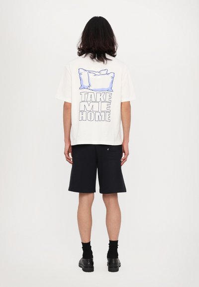 T-shirt bianco di cotone con una grafica raffigurante due cuscini sopra il testo "PORTAMI A CASA" in lettere in grassetto. Abbinato a pantaloni corti blu navy.