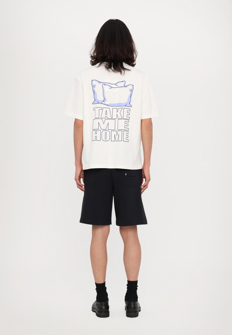 Witte katoenen t-shirt met een grafisch ontwerp van twee kussens boven de tekst "NEEM ME MEE NAAR HUIS" in vette letters. Gecombineerd met marineblauwe shorts.