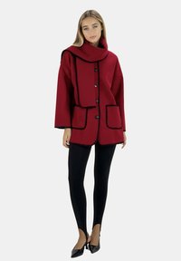 Abrigo de lana rojo con detalles en negro, cierre de botones, grandes bolsillos y cuello de chal, combinado con leggings negros y tacones.