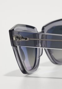 Pilki permatomi akiniai nuo saulės, turintys geometrinį dizainą, sidabro metalinius akcentus ir "Ray-Ban" logotipą ant rėmelio.