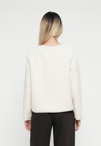 Pull en polaire texturé de couleur crème, avec un col rond, des manches longues et une coupe décontractée, associé à un pantalon sombre.