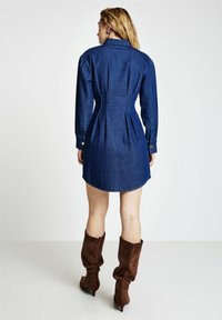 Jeanshemdkleid in Dunkelblau mit langen Ärmeln, tailliertem Schnitt und Falten am Rücken. Kombiniert mit braunen, lässigen kniehohen Stiefeln.