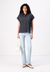 Pelēka īsām piedurknēm krekls ar divām krūšu kabatām, apvienots ar gaiši zilu platā stila džinsu biksēm un taupes krāsas apaviem, tur rokā baltu somu.