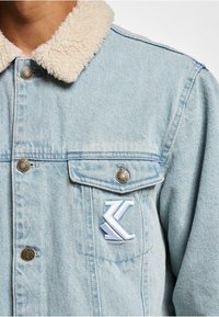 Světle modrá denimová bunda s měkkým, béžovým beránkovým límcem, která má stříbrné knoflíky a bílou vyšívanou logo na hrudníku.