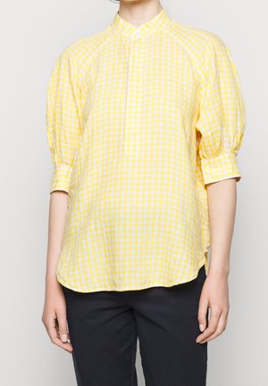 Persoon die een geel-wit geruite blouse met driekwart pofmouwen en een zwarte broek draagt tegen een effen achtergrond.