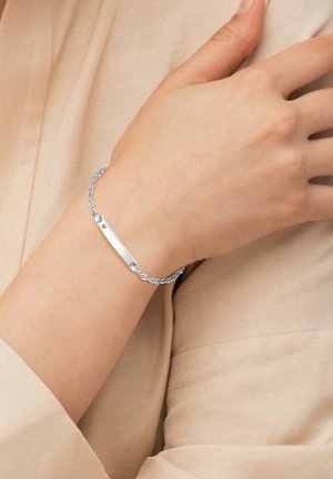 Bracciale d'argento con un design a catena intrecciata e una piastra liscia e allungata con un piccolo cuore come accento. Indossato su un polso contro un tessuto chiaro.