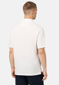 Bílá polo košile s krátkým rukávem, která má klasický límec, boční rozparky a jemnou texturu, s hladkým povrchem a minimalistickým designem.