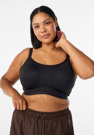 Žena s čiernymi vlasmi po ramená, oblečená v čiernu športovú podprsenku a hnedé nohavice so šnúrkou, dotýkajúca sa krku a pásu na bielom pozadí.