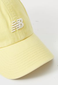Šviesiai geltona beisbolo kepuraitė su baltu siuvinėjamu New Balance logotipu priekyje, prieš baltą foną.