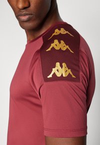 Bordeaux sportshirt met korte mouwen, voorzien van een gestructureerd bordeaux schouderpaneel en gouden logo-accenten. Gladde stof met een aangesloten ontwerp.
