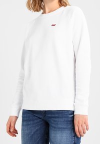 Sudadera blanca con cuello redondo, mangas raglán y un pequeño logo rojo en el pecho. Fabricada en algodón, cuenta con puños y dobladillo acanalados.