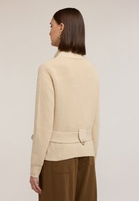 Béžový pletený svetr s vysokým límcem, žebrovanou strukturou a detailem v pase se zavazováním. Má dlouhé rukávy a volný střih.