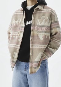 Giacca camicia in tessuto a tonalità pastello con motivi geometrici, indossata sopra una felpa nera con cappuccio con testo del logo bianco, abbinata a jeans blu.