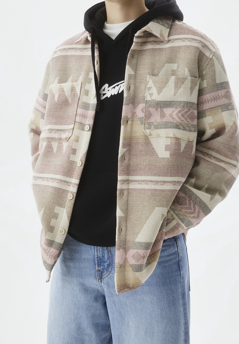Giacca camicia in tessuto a tonalità pastello con motivi geometrici, indossata sopra una felpa nera con cappuccio con testo del logo bianco, abbinata a jeans blu.