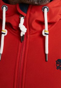 Veste rouge avec une fermeture éclair à l'avant, comprenant des cordons blancs avec des accents colorés et un logo brodé sur la poitrine.